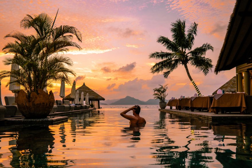 Guest in an infinity pool at sunset beside palms and a beachfront restaurant